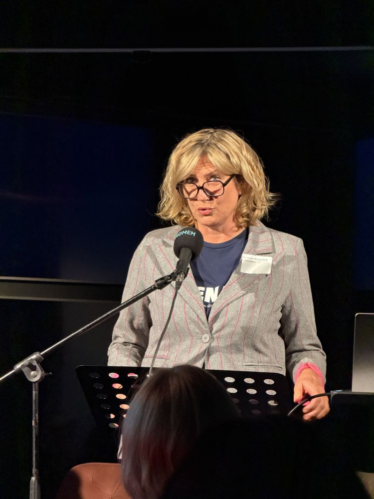 Christiane Wiesenfeldt speaking into a microphone at MOMEM in Frankfurt, wearing glasses and a grey blazer, during her presentation on techno and historical musicology.